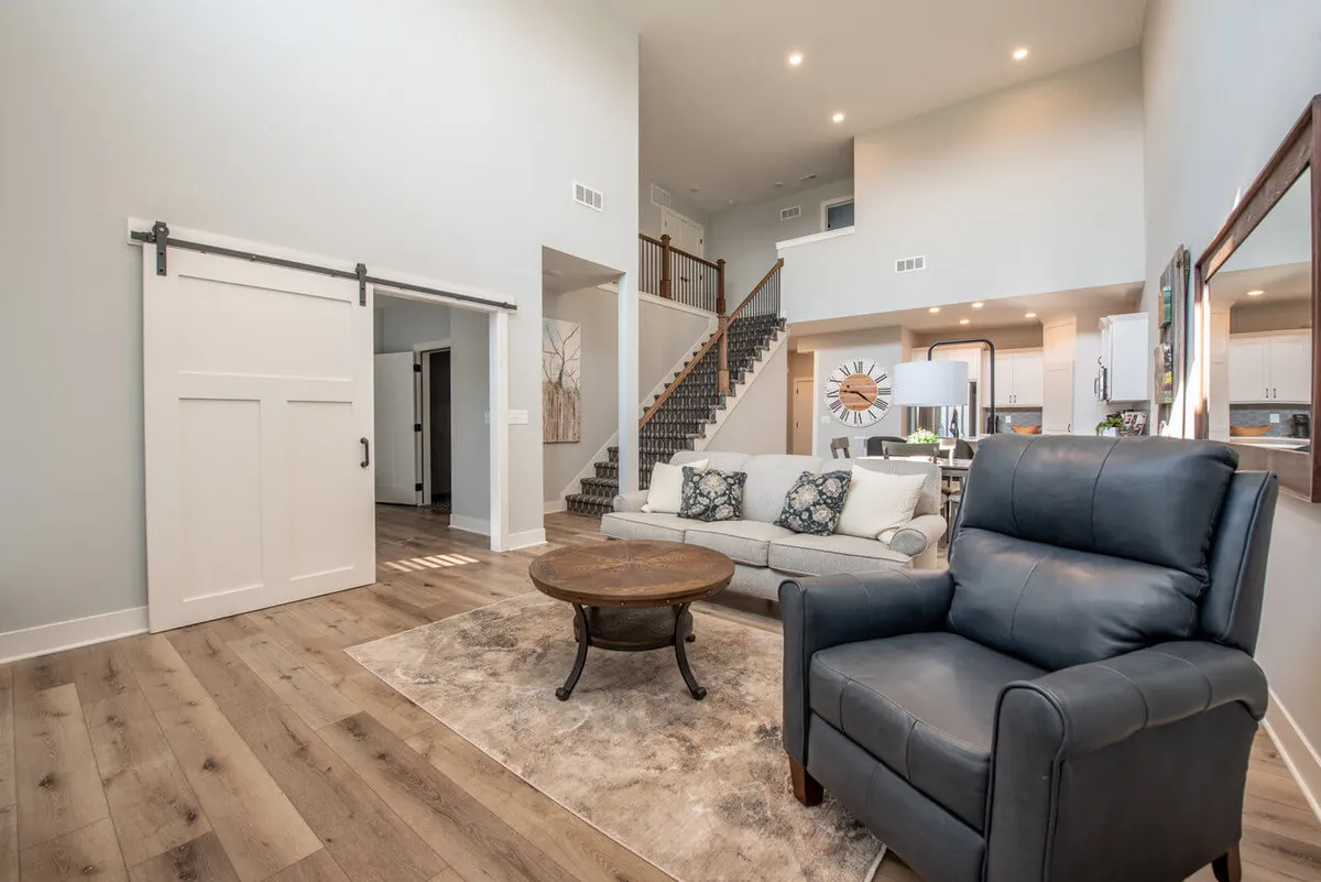 A spacious, open-plan living area featuring a comfortable sofa, a sleek coffee table, a visible staircase, and a stylish sliding barn door. The room is designed for relaxing and socializing, with ample space and an inviting atmosphere.