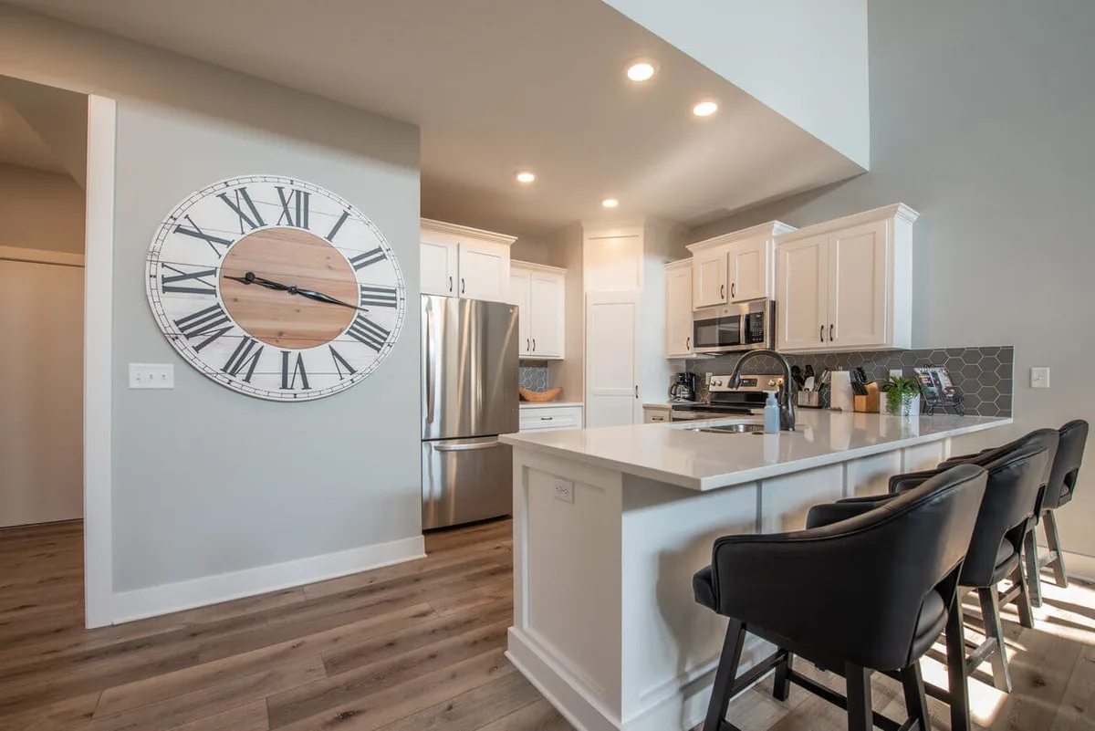 A modern kitchen scene with a prominent kitchen island bordered by bar stools, stainless steel appliances, and a large wall clock. The open space invites casual dining, conversation, and culinary creativity.