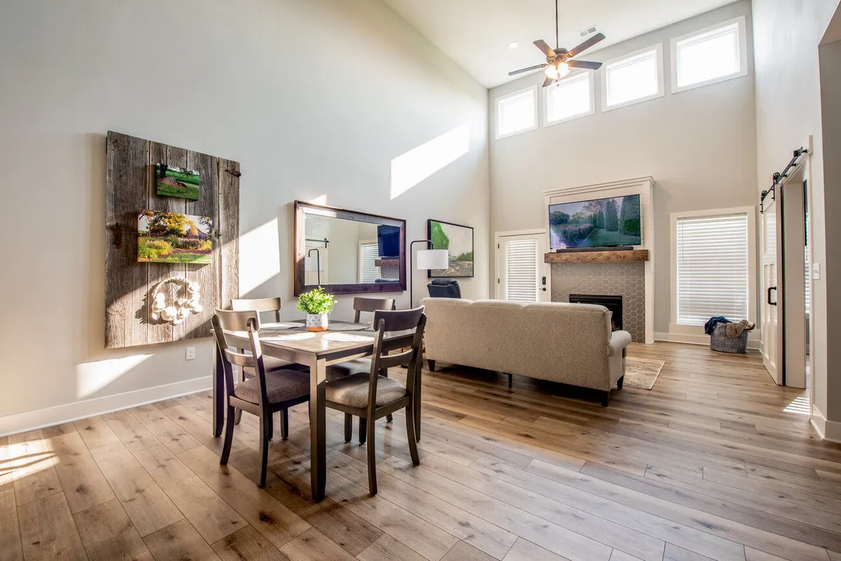 An open-concept living and dining area with a large sofa, dining table, tasteful wall art, and prominent large windows. The space feels warm and inclusive, ideal for family meals, socializing, or relaxing with coffee and conversation.