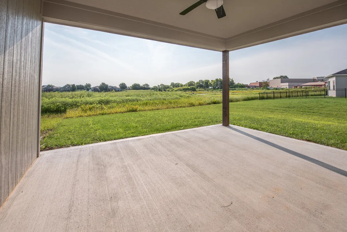 A tranquil open patio area overlooks a lush green field, framed by distant trees under a bright sky. The patio provides ample space for outdoor dining, relaxation, or gatherings, while the adjacent grassy expanse evokes a sense of connection to the outdoors and natural beauty.