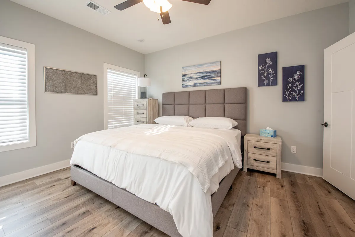A serene bedroom featuring a gray upholstered headboard, a neatly made bed, a nightstand with a softly glowing lamp, and decorative wall art. Natural light gently filters through a nearby window, creating a peaceful and inviting atmosphere ideal for rest.