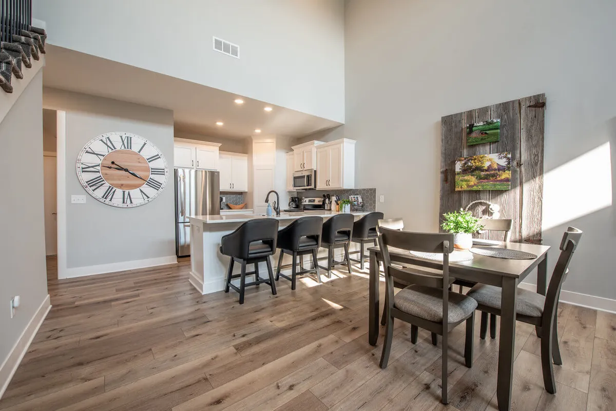 An inviting combined kitchen and dining area with a large wall clock, kitchen island, a spacious dining table surrounded by chairs, and decorative wall art. The open arrangement provides easy access and plenty of space for group dining or special occasions.